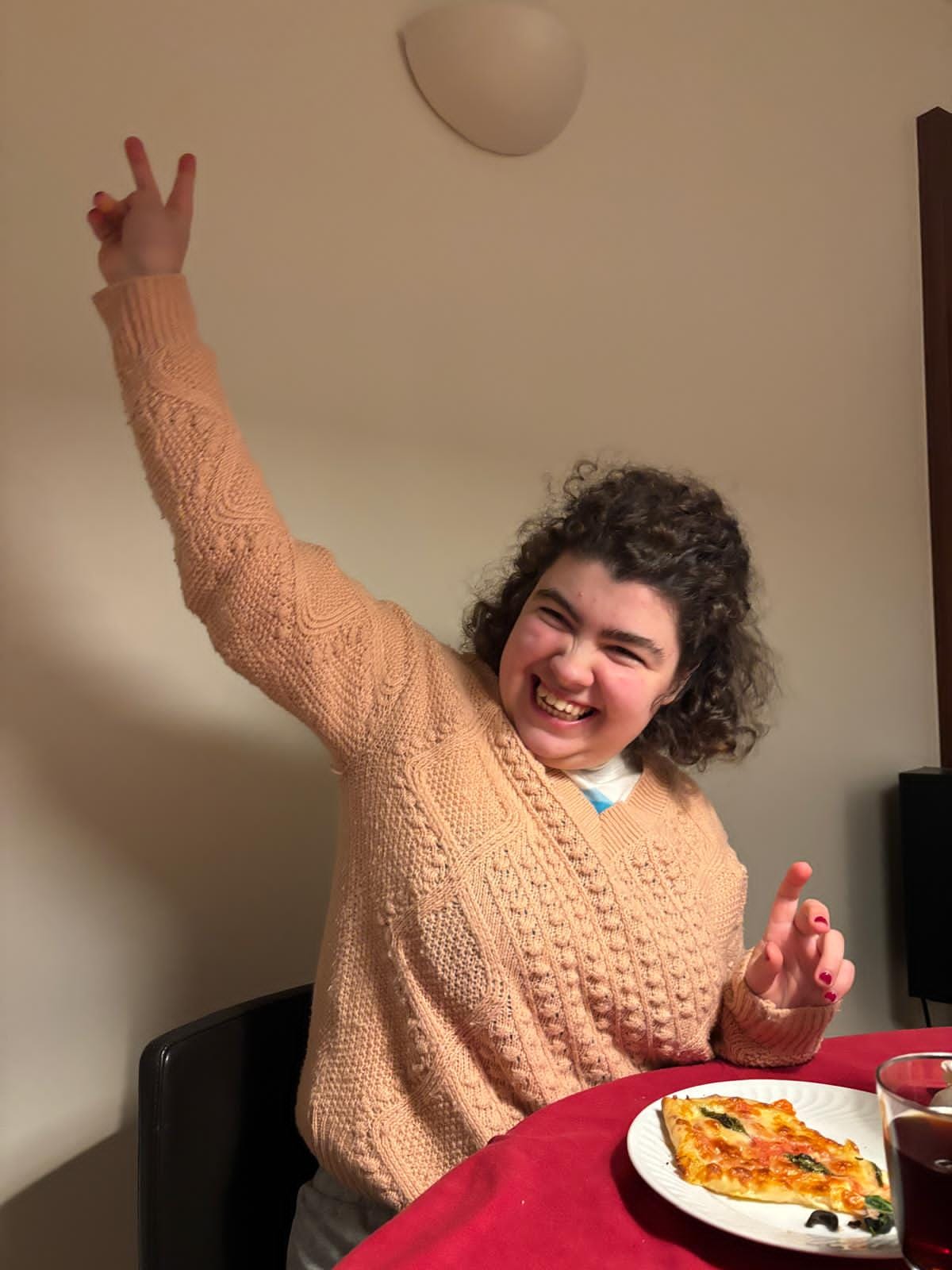 Leonor, uma jovem adulta, à mesa com uma fatia de pizza, a fazer o sinal de vitória
