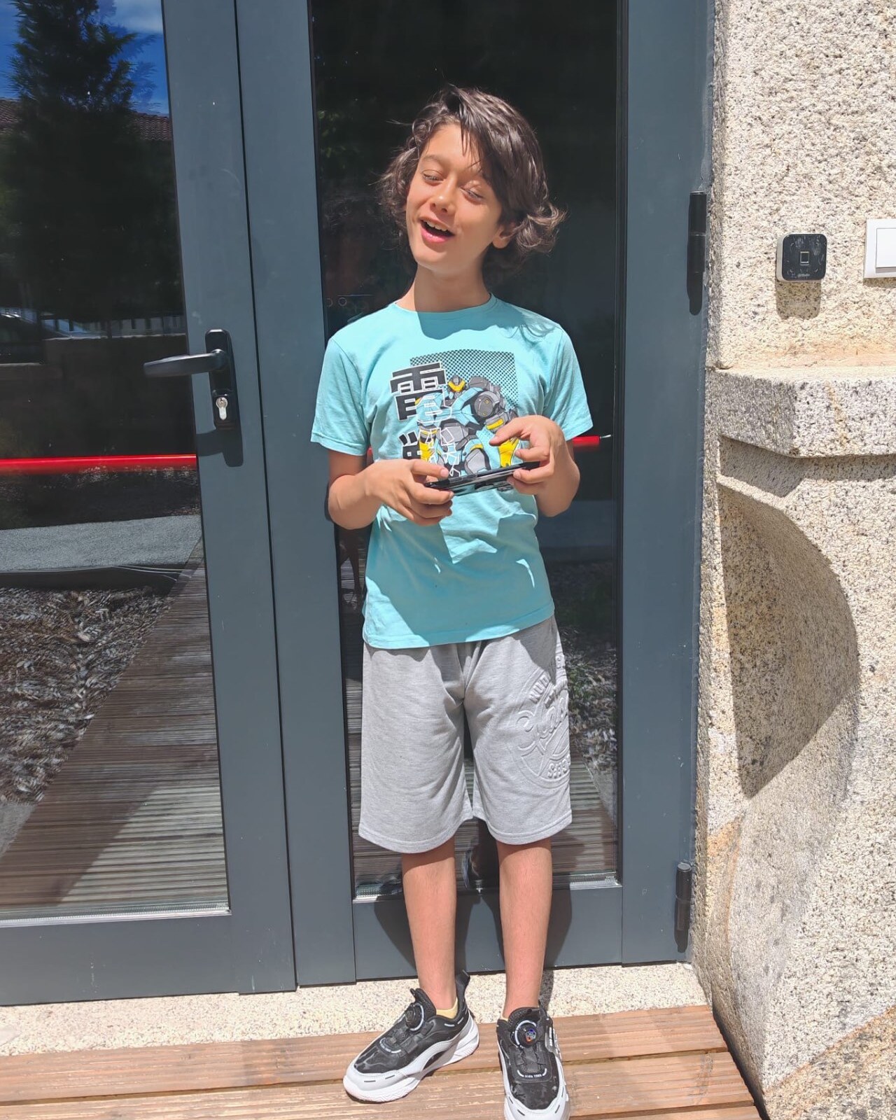 Rafael, um rapaz de 12 anos, de t-shirt azul, a segurar um telemóvel à porta de um prédio