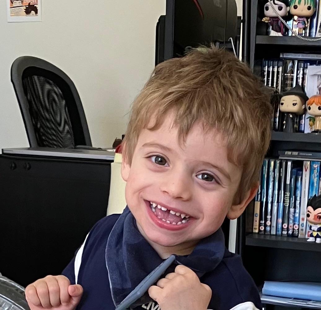 Rafa, um menino de 3 anos, a sorrir em frente a uma estante com livros e figuras